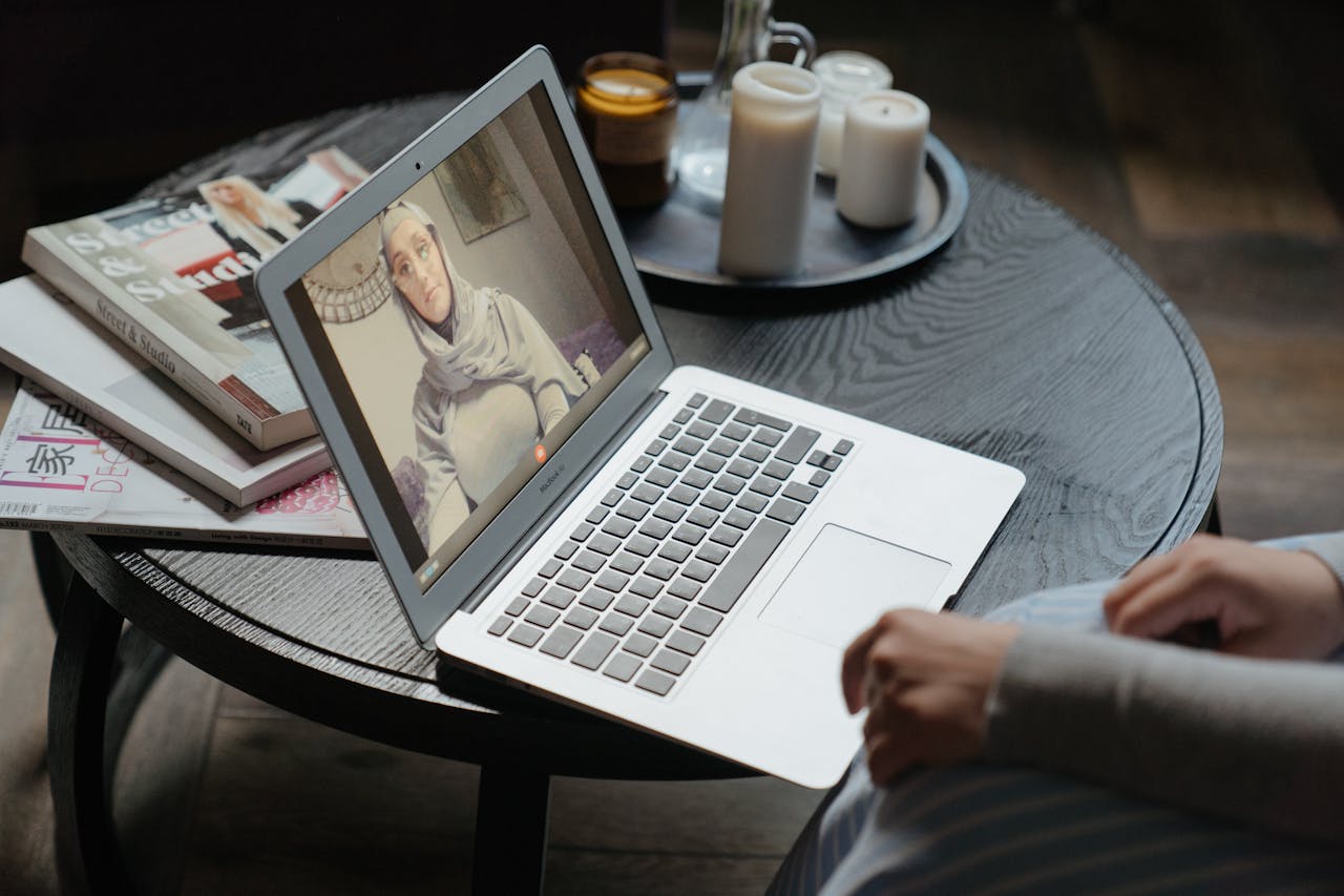 A woman in a hijab participating in a video call on a laptop, seated at a round table indoors.